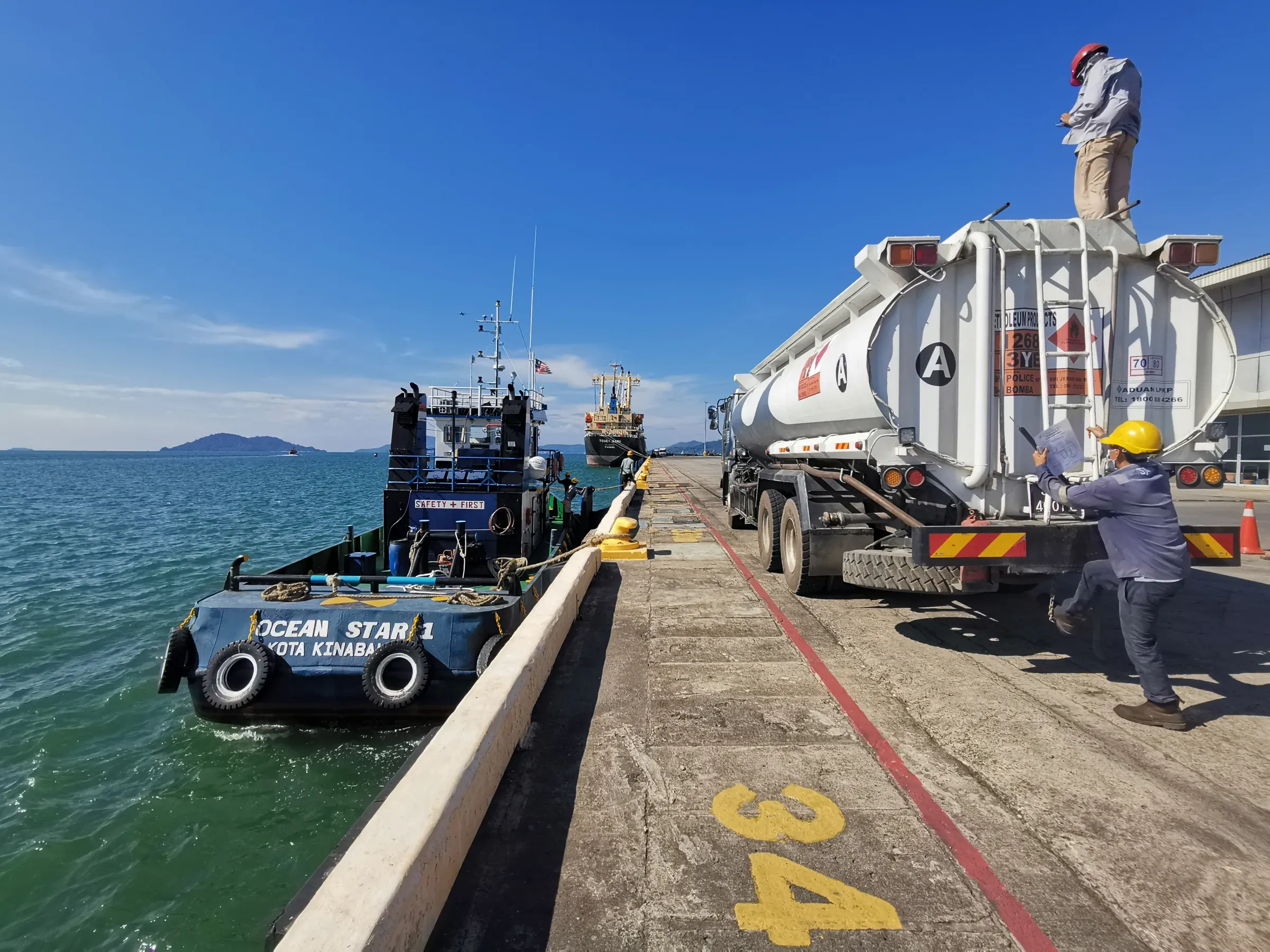 A tug and a tanker truck at the quay, with a worker on top of the truck